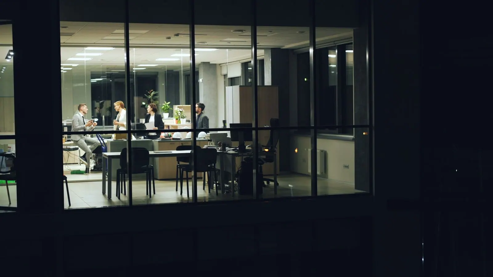 An office lit at night, seen through its glass facade, with a team still at work